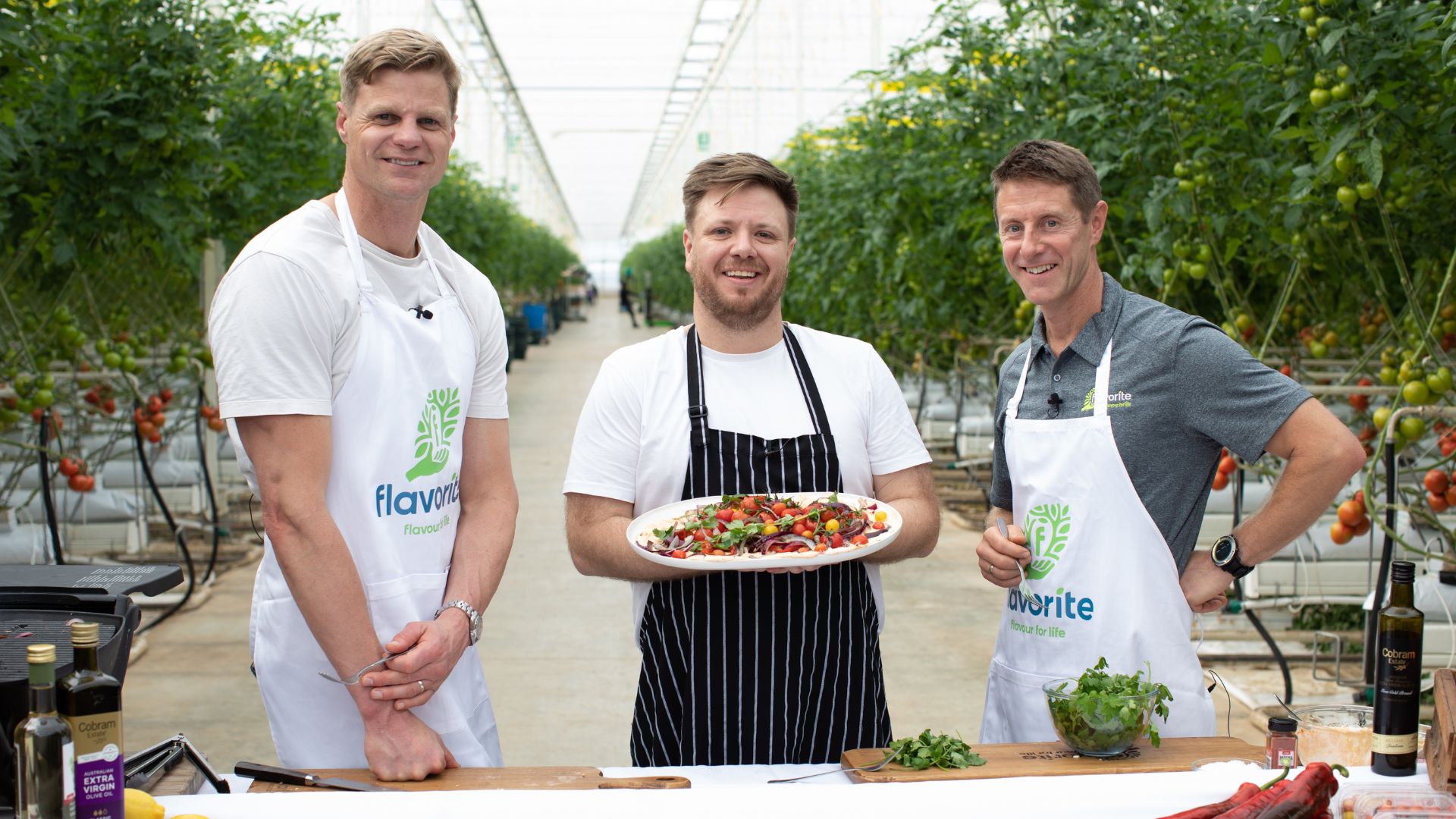 In a glasshouse, Nick Riewoldt stands with Michael Weldon and Chris Millis. Michael is holding a plate of fresh produce.